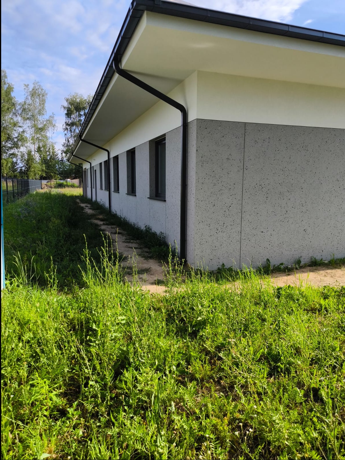 Nowoczesny budynek z szarą elewacją typu baranek i białym pasem pod dachem, widok z poziomu trawy