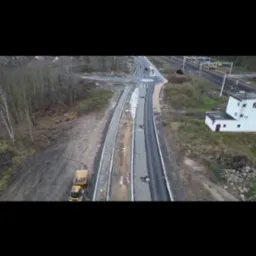 Widok z góry na budowę drogi z nowym asfaltem i żółtą ciężarówką wywożącą ziemię, w tle drzewa i budynek