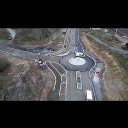 BUDMET - Zdjęcie z drona przedstawia budowę ronda z widocznymi maszynami budowlanymi, robotnikami w pomarańczowych kamizelkach i świeżo położonym asfaltem. W tle widoczne tereny zielone i częściowo...