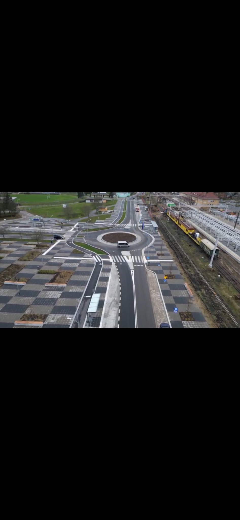 Widok z lotu ptaka na rondo z kostki brukowej w szachownicę, obok torów kolejowych z żółtym pociągiem. Na rondzie stoi srebrny samochód, a w tle widać zielony teren.