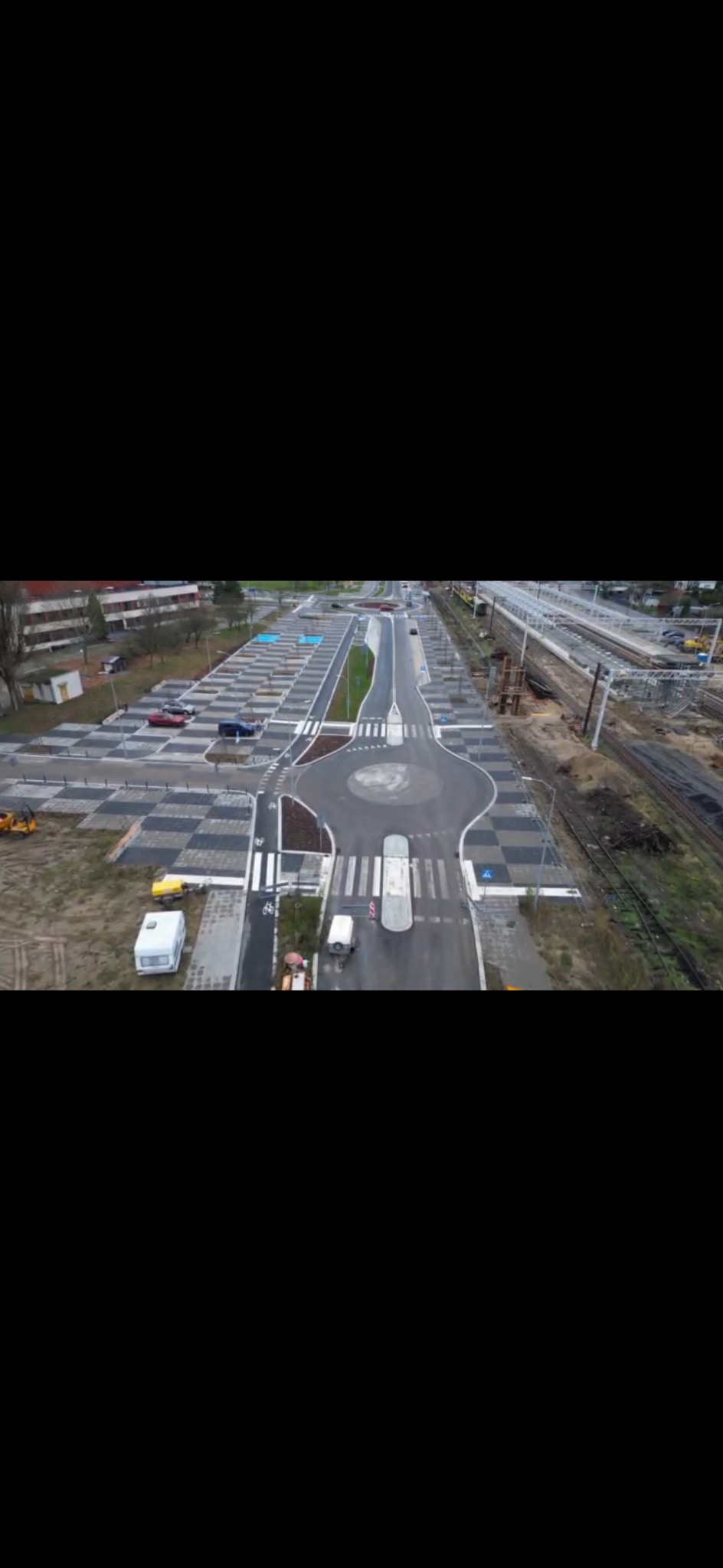 Widok z lotu ptaka na nowo wybudowane rondo z kostki brukowej i parking z szachownicą z kostki, obok torów kolejowych i budynków mieszkalnych.