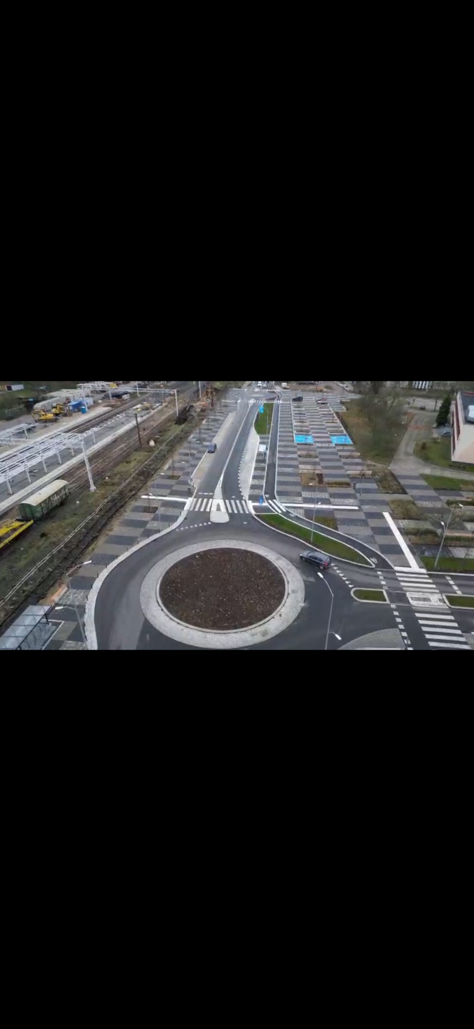 Widok z góry na nowo wybudowane rondo z kostki brukowej, z czarnym samochodem na nim, w tle tory kolejowe i parking z kostki brukowej w szachownicę.