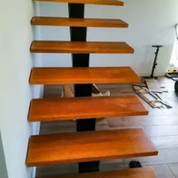 Ażurowe schody z drewnianymi stopniami o ciepłym odcieniu i czarną, metalową konstrukcją wspornikową, widoczne z dołu, z elementami kartonu i sprzętu czyszczącego w tle.