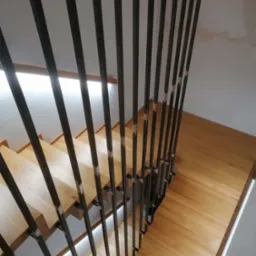Drewniane schody z nowoczesną balustradą z pionowych, metalowych prętów, widok z góry na stopnie oświetlone od spodu.