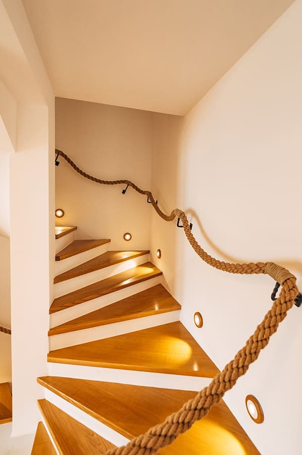 Spiralne schody z drewnianymi stopniami, oświetleniem punktowym i balustradą z grubego liny jutowej, mocowanej do ściany za pomocą czarnych uchwytów.