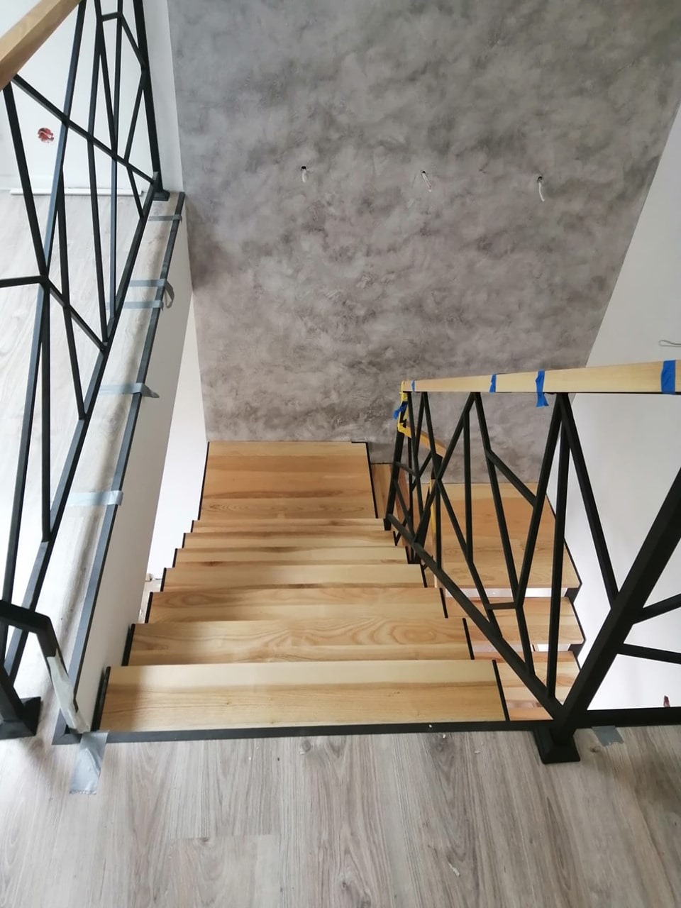 Widok z góry na jasne, drewniane stopnie schodów z czarną, geometryczną balustradą w Kętach, częściowo zabezpieczoną taśmą malarską, na tle szarej, dekoracyjnej ściany.