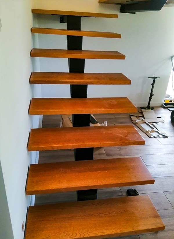 Ażurowe schody z drewnianymi stopniami o ciepłym odcieniu i czarną, metalową konstrukcją wspornikową, widoczne z dołu, z elementami kartonu i sprzętu czyszczącego w tle.