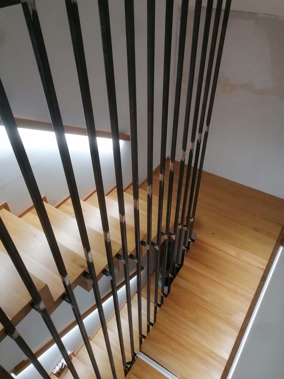 Drewniane schody z nowoczesną balustradą z pionowych, metalowych prętów, widok z góry na stopnie oświetlone od spodu.