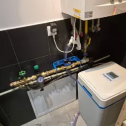 Instalacja uzdatniania wody Aquaphor w pomieszczeniu z czarnymi płytkami, widoczne rury miedziane, zawory z zielonymi pokrętłami i manometr. Nad instalacją piec gazowy z żółtą rurą doprowadzającą gaz.