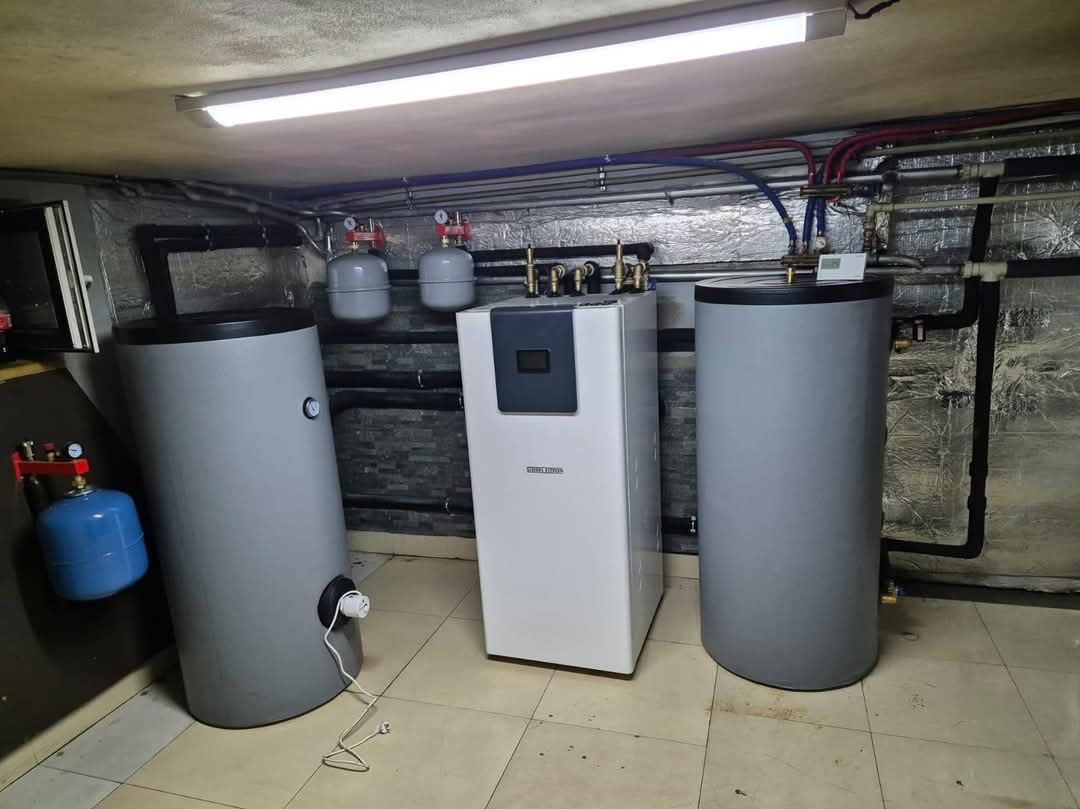 Biała pompa ciepła Steibel Eltron pomiędzy dwoma szarymi zasobnikami wody w piwnicy z rurami na suficie i ścianie wyłożonej folią aluminiową.