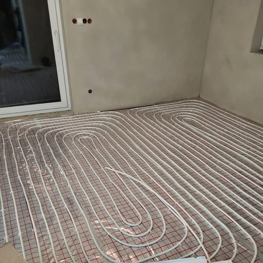 Białe rury ogrzewania podłogowego ułożone na czerwonej siatce w surowym wnętrzu z oknem i szarą ścianą.