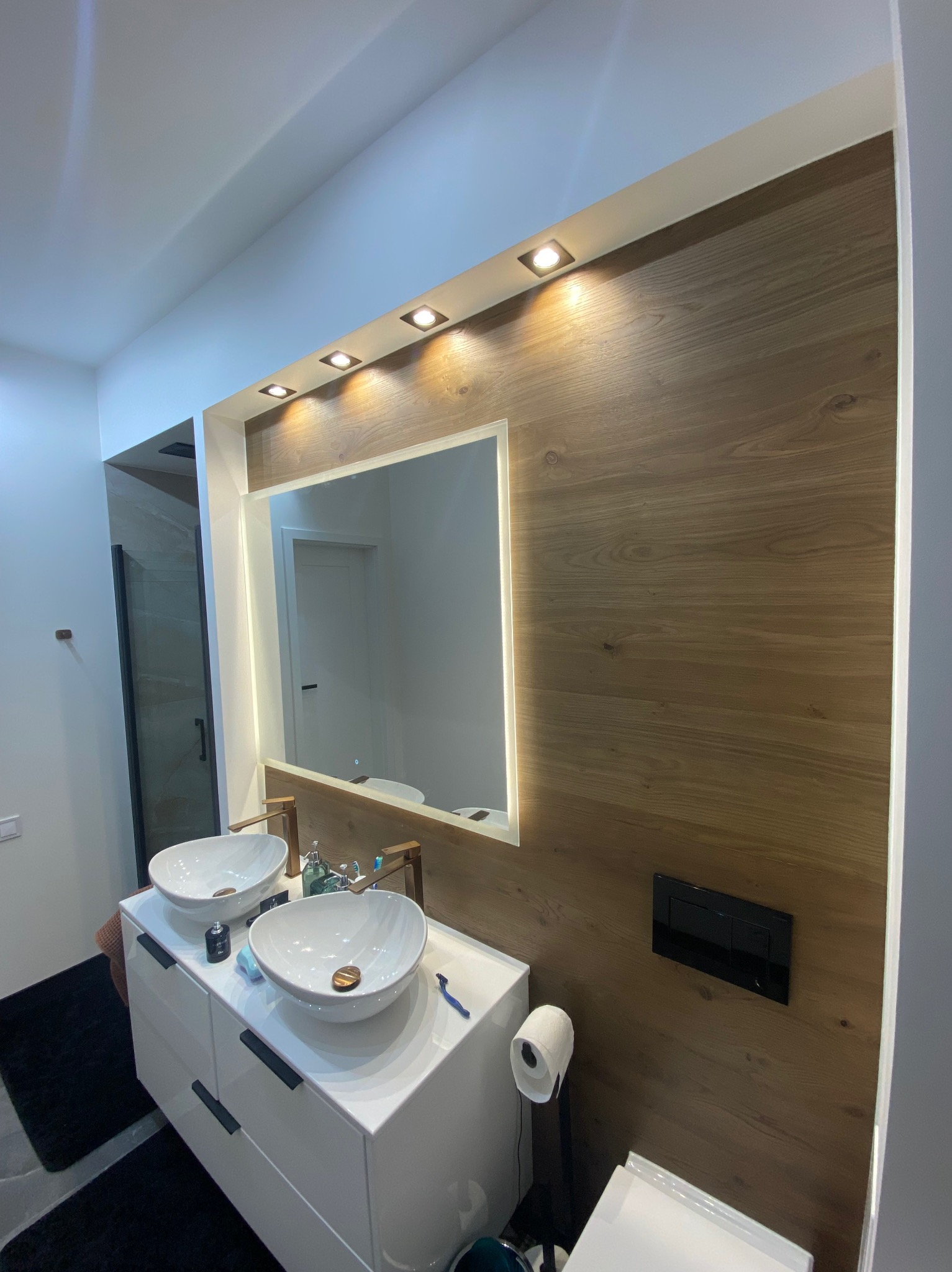 Nowoczesna łazienka z drewnianą ścianą, podświetlanym lustrem i dwiema umywalkami nablatowymi na białej szafce. Widoczna toaleta i czarny dywanik.