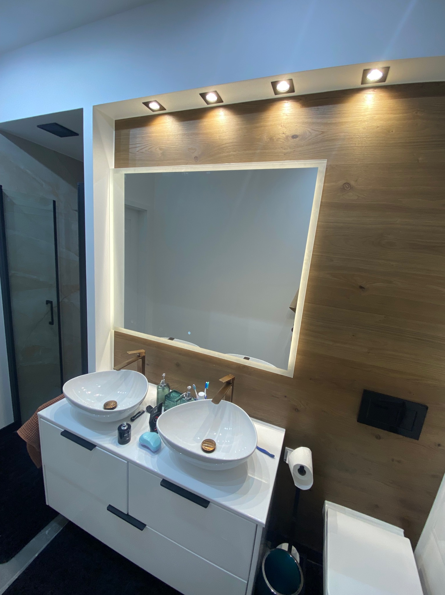 Nowoczesna łazienka z dwiema umywalkami nablatowymi, drewnianą ścianą i podświetlanym lustrem. Widoczna kabina prysznicowa w tle.