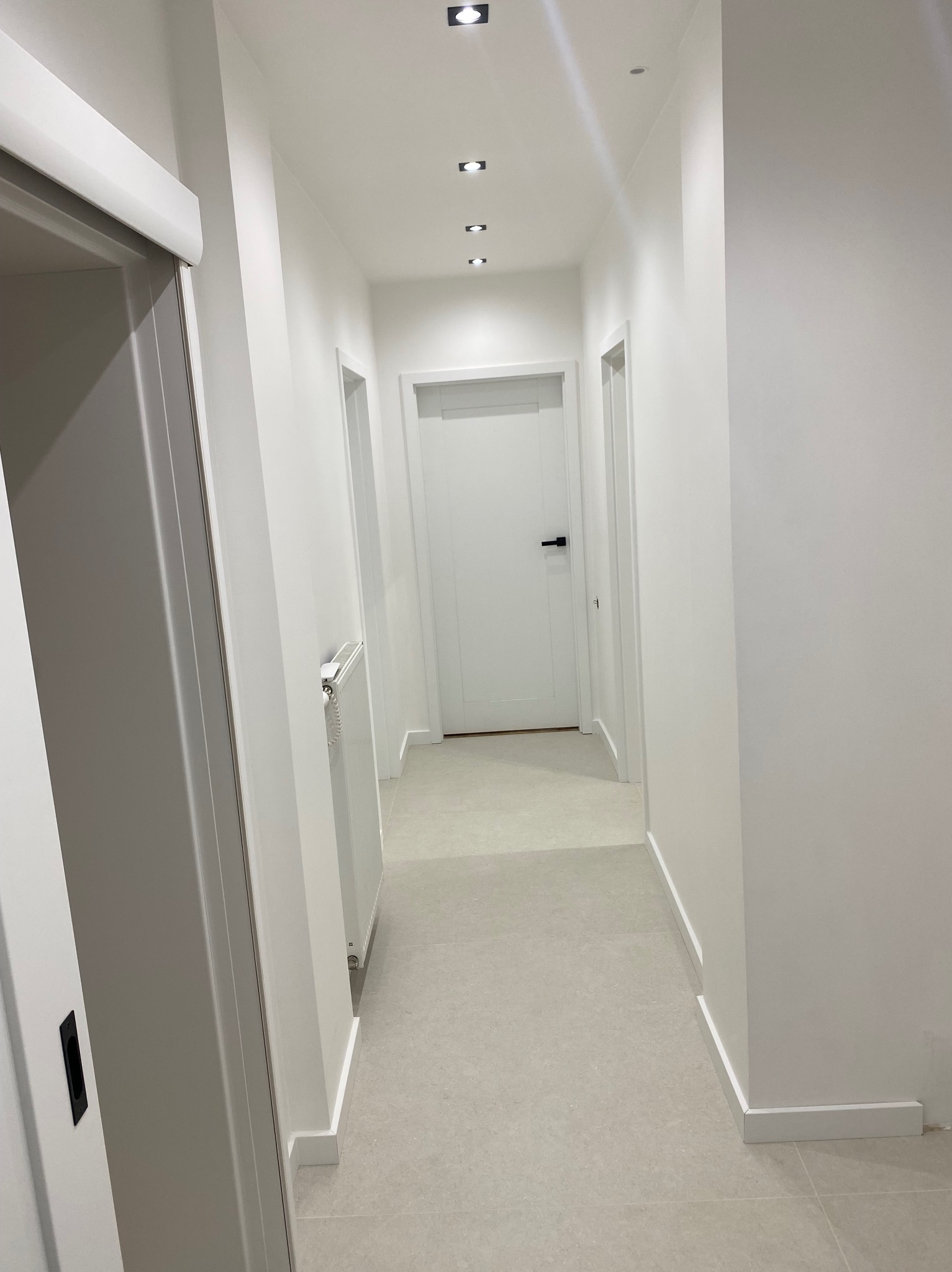 Biały korytarz po remoncie z szarymi płytkami podłogowymi i minimalistycznym oświetleniem sufitowym. Widoczne drzwi i grzejnik.
