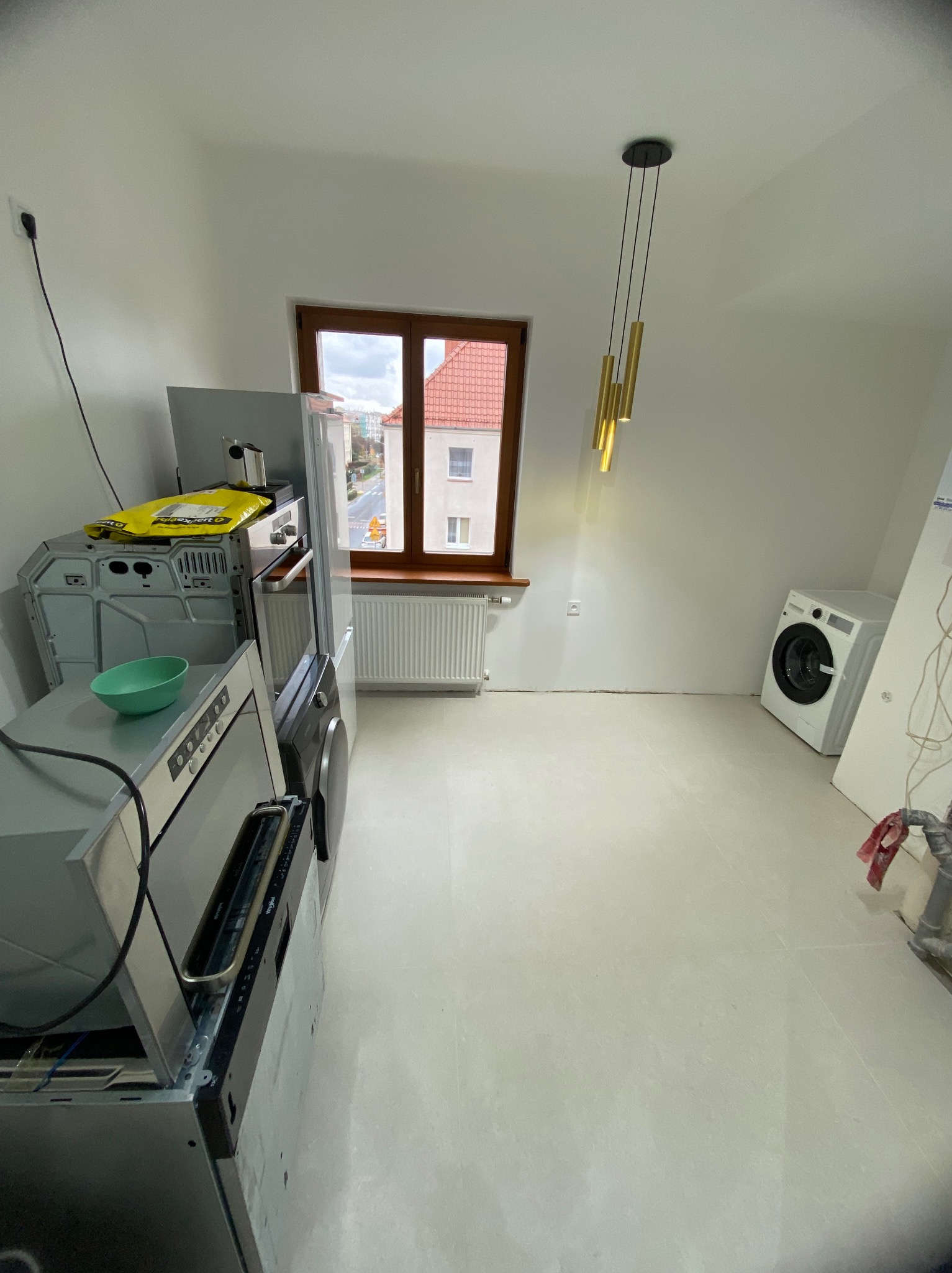 Wnętrze w trakcie wykończenia: białe ściany i podłoga, okno z widokiem na miasto, sprzęt AGD częściowo rozpakowany, lampa wisząca. Przygotowanie do montażu.
