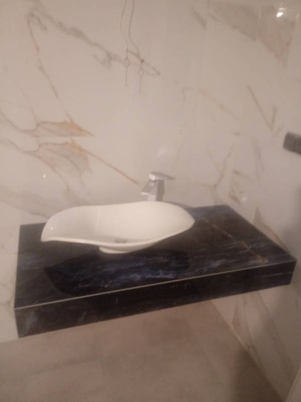 Umywalka nablatowa o nietypowym kształcie, zamontowana na blacie z ciemnego marmuru z niebieskimi żyłkami, ściana wyłożona płytkami imitującymi marmur z jasnymi żyłkami, widoczne niedokończone...