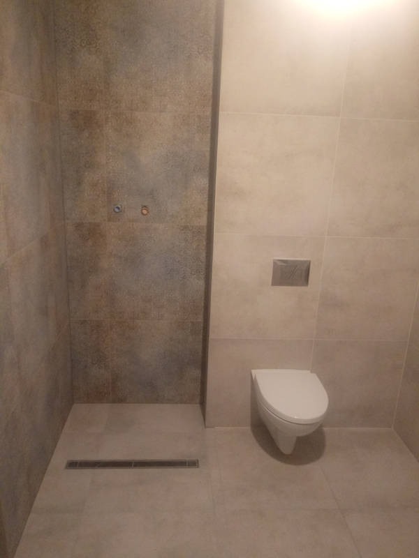 Wykończona łazienka z beżowymi płytkami, widoczny odpływ liniowy w strefie prysznicowej i podwieszana toaleta z chromowanym przyciskiem spłukującym.