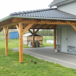 Drewniana wiata garażowa z ciemną dachówką ceramiczną, połączona z budynkiem, z widocznymi rynnami i kostką brukową na podjeździe.