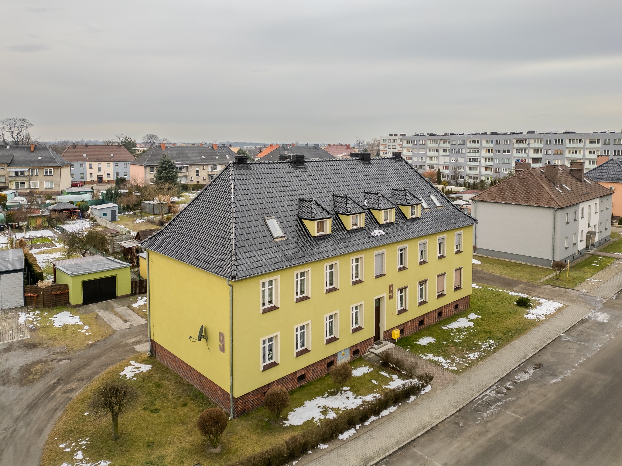 Widok z góry na dwupiętrowy, żółty budynek mieszkalny z nowym, czarnym dachem z dachówki ceramicznej i oknami dachowymi, w tle inne budynki i pochmurne niebo.