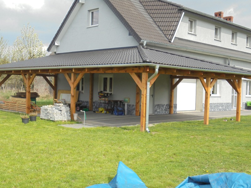 Zadaszenie tarasu z drewnianą konstrukcją i grafitową dachówką ceramiczną, widoczne rynny w kolorze srebrnym, na trawniku materiały budowlane i narzędzia.