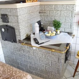 Piecyk - Drelinkiewicz Adam - Kuchenny piec kaflowy z granitowych płytek, z czarnymi drzwiczkami i blatem, na którym leżą jabłka w misce, pilot do telewizora, nóż i butelki z płynami.