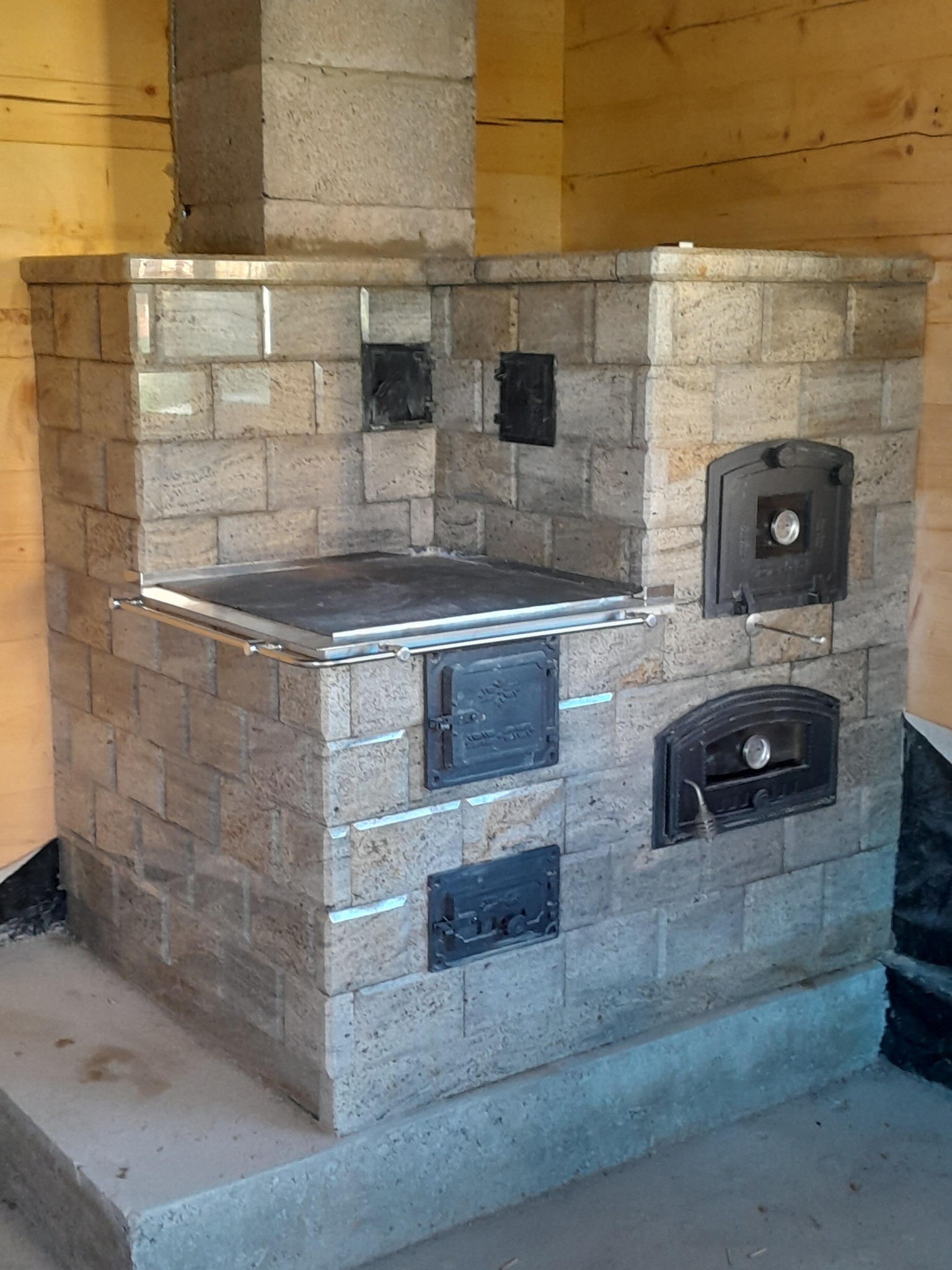 Kuchenny piec kaflowy z jasnych, granitowych kafli, z żeliwną płytą grzewczą i metalowymi drzwiczkami paleniska oraz piekarnika, umieszczony w drewnianej wnęce.