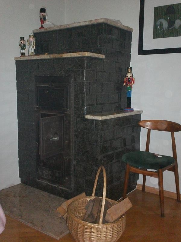 Czarny piec kaflowy z otwartymi drzwiczkami, wykończony marmurowymi półkami, ozdobiony figurkami dziadków do orzechów, obok stoi wiklinowy kosz z drewnem i krzesło.