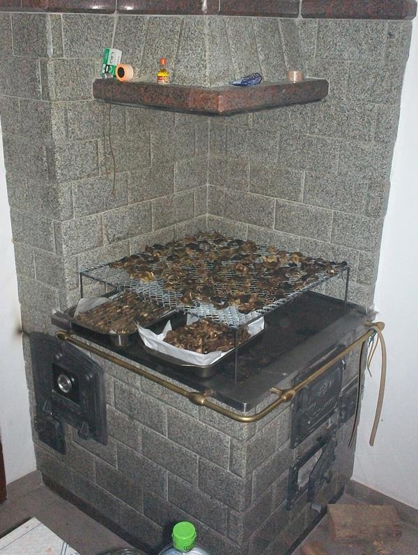 Granitowy piec kuchenny z suszonymi grzybami na ruszcie i w blachach, widoczne drzwiczki paleniskowe i półka z drobiazgami.