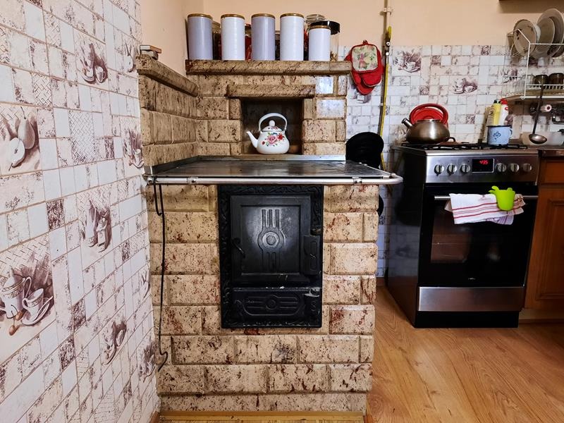 Kuchenny piec kaflowy z czarnymi drzwiczkami i metalowym blatem, otoczony płytkami z motywem owoców i naczyń, w tle widoczna kuchenka gazowa.
