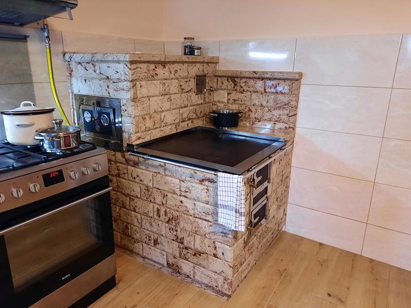 Kaflowa kuchnia z piekarnikiem, płytą grzewczą i dolnymi drzwiczkami, obok stoi kuchenka gazowa z garnkami, całość w jasnej kuchni z drewnianą podłogą.