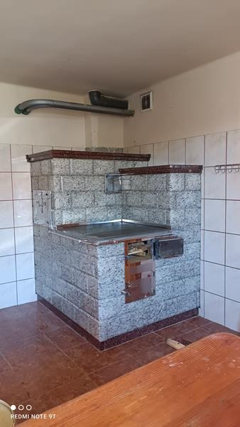 Kuchenna zabudowa pieca kaflowego z szarych płytek imitujących granit, z metalowym blatem i drzwiczkami, widoczna rura odprowadzająca dym.