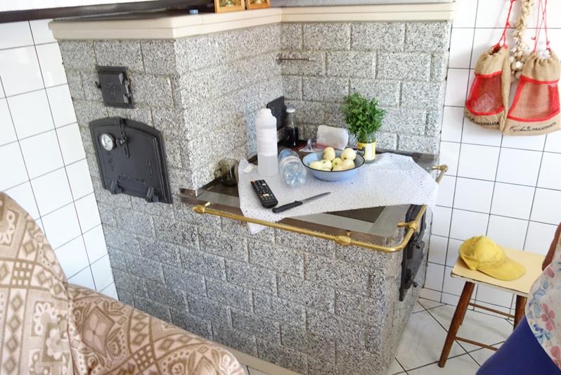 Kuchenny piec kaflowy z granitowych płytek, z czarnymi drzwiczkami i blatem, na którym leżą jabłka w misce, pilot do telewizora, nóż i butelki z płynami.