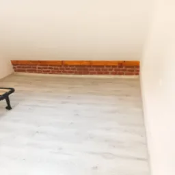 Poddasze z jasnymi panelami podłogowymi i dekoracyjnym fragmentem ceglanej ściany pod skosem dachu; widoczny fragment ramy łóżka.