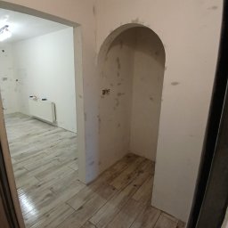 SANITREM Stanislaw Medvediuk - Wnętrze w trakcie prac wykończeniowych: ułożona podłoga z płytek imitujących drewno, ściany przygotowane pod malowanie, wnęka z łukiem. Widok z perspektywy wejścia.