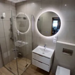 Nowoczesna łazienka z kabiną prysznicową typu walk-in, okrągłym lustrem z podświetleniem LED i podwieszaną szafką z umywalką. Płytki imitujące drewno na podłodze.