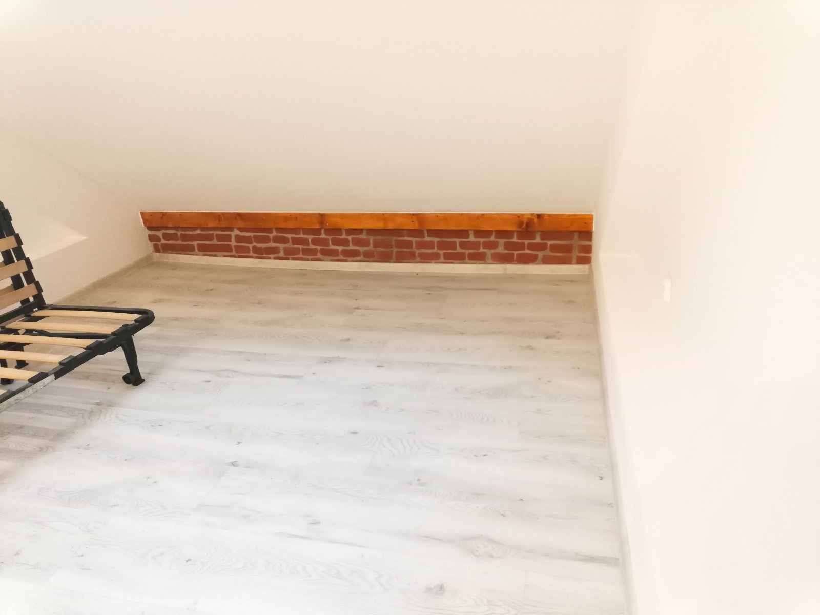 Poddasze z jasnymi panelami podłogowymi i dekoracyjnym fragmentem ceglanej ściany pod skosem dachu; widoczny fragment ramy łóżka.