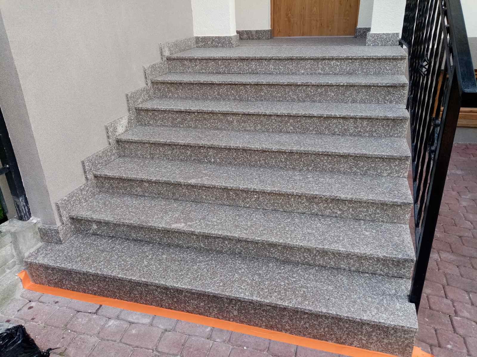 Schody zewnętrzne wyłożone szarym, nakrapianym kamieniem, z widocznymi fragmentami czarnej metalowej balustrady i pomarańczowego zabezpieczenia na dole