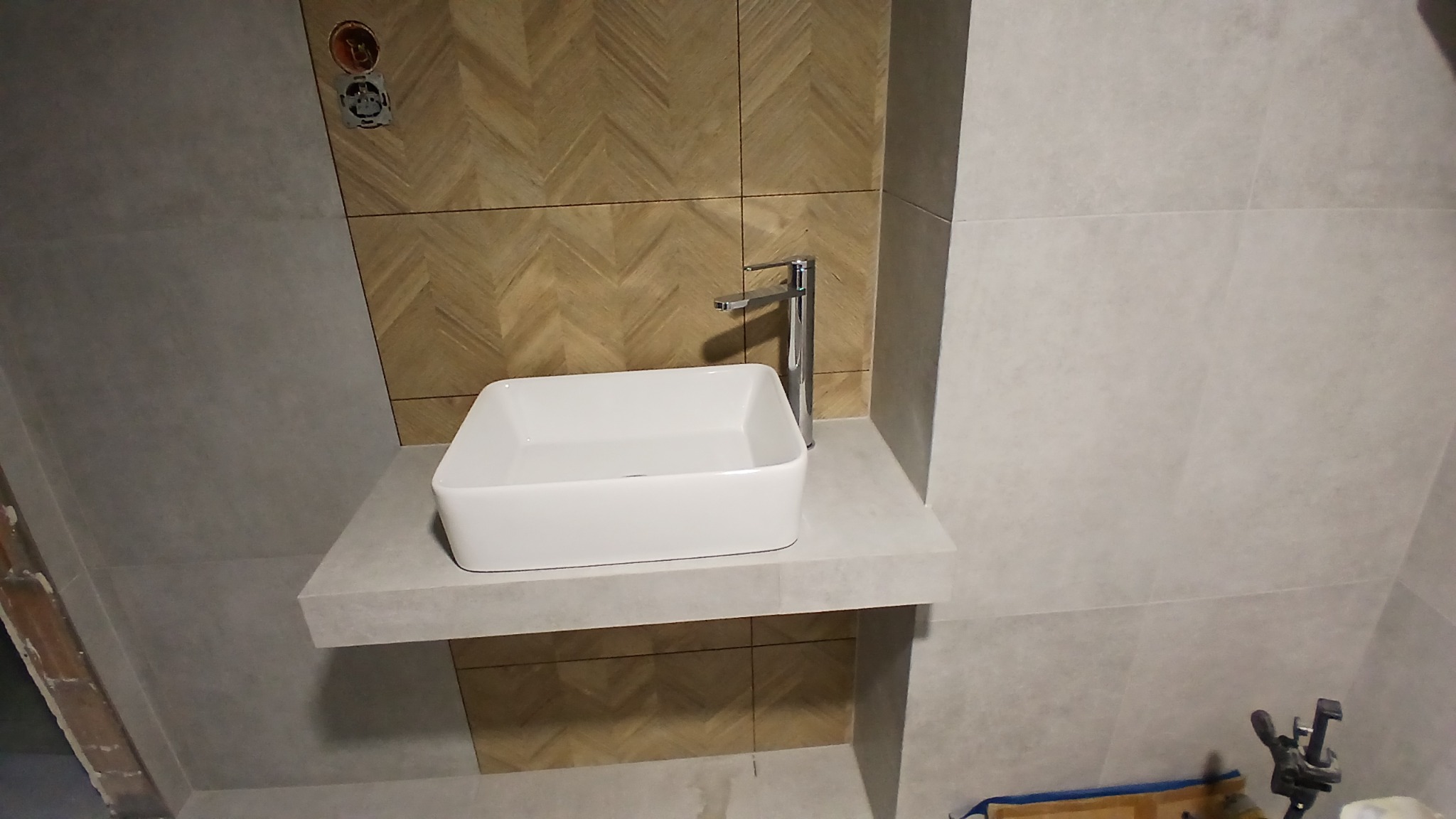 Nowoczesna łazienka z umywalką nablatową na szarym blacie. Na ścianie płytki w geometryczny wzór i szare płytki. Srebrna bateria. Widoczne narzędzia w dolnym rogu.