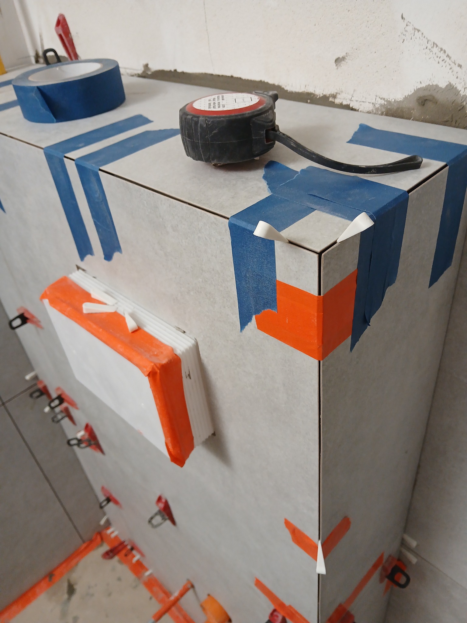 Szare płytki na zabudowie, zabezpieczone niebieską taśmą malarską, z użyciem pomarańczowych klinów i klipsów poziomujących. Na górze miara i rolka taśmy.