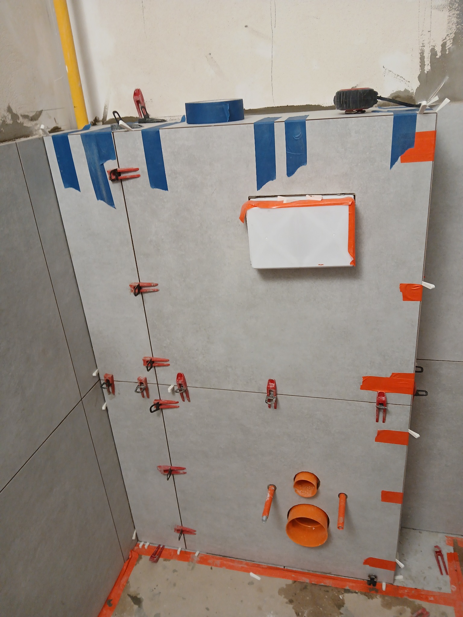 Montaż płytek w łazience z użyciem klipsów poziomujących i taśmy maskującej. Widoczne otwory na instalacje wod-kan. Etap prac wykończeniowych.