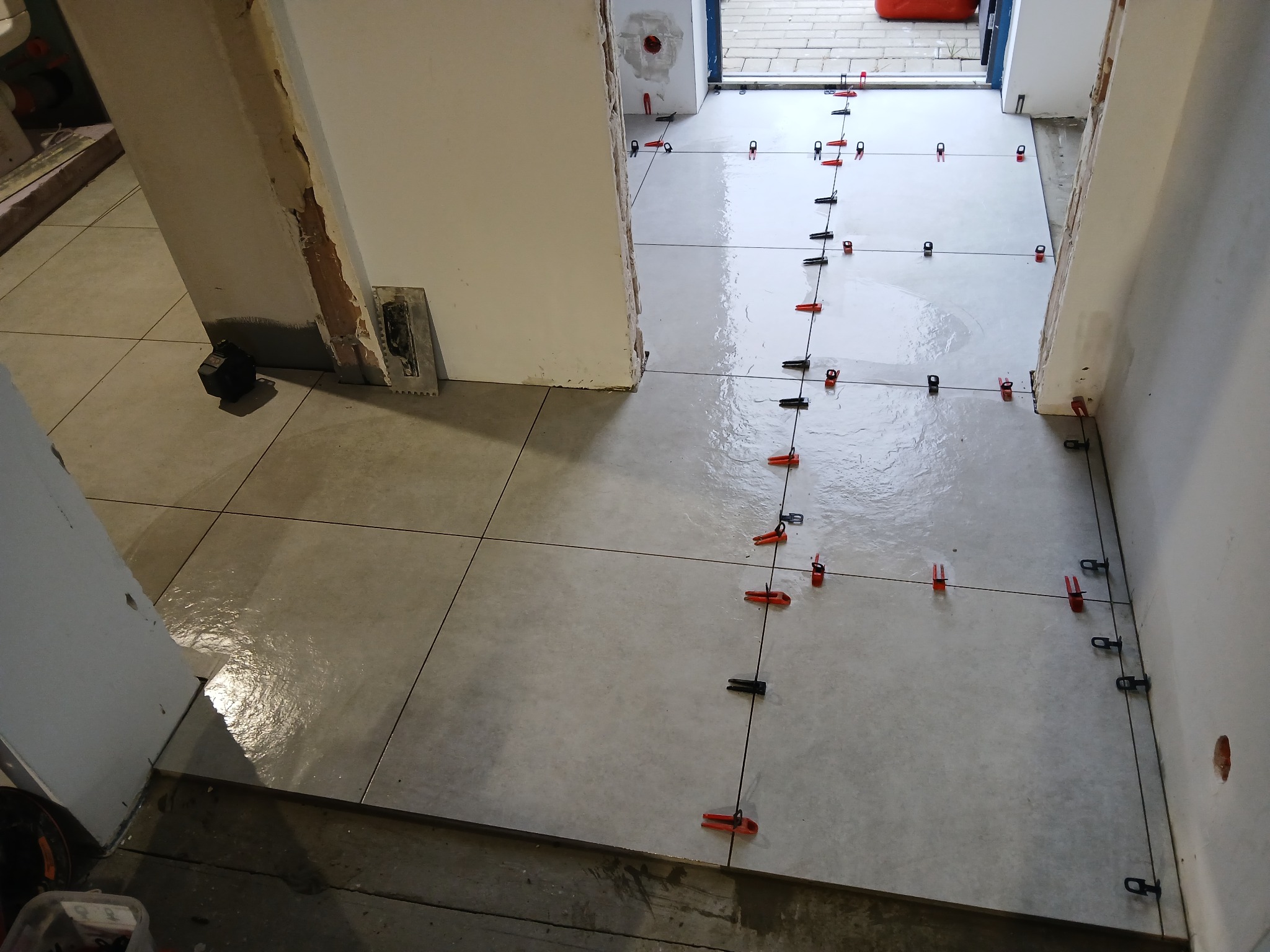 Świeżo ułożone, jasne płytki podłogowe z systemem poziomowania, widoczne fugi i narzędzia glazurnicze przy ścianie. Prace w trakcie wykańczania.