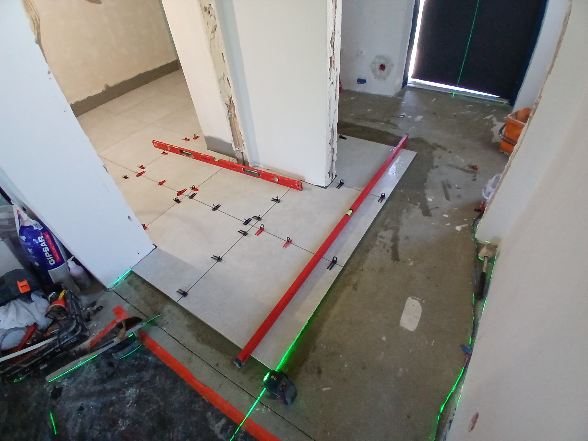 Układanie jasnych płytek podłogowych z użyciem poziomicy laserowej i krzyżyków dystansowych. Widoczne narzędzia glazurnicze i poziomice. Prace w toku.