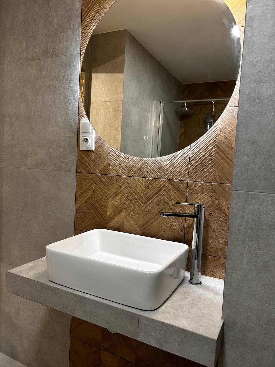Nowoczesna łazienka po remoncie: biała, prostokątna umywalka nablatowa, chromowana bateria, okrągłe lustro z podświetleniem i dekoracyjna ściana z drewnopodobnych płytek.