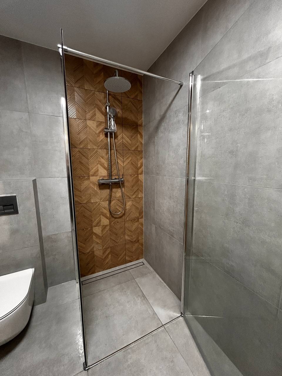 Nowoczesna kabina prysznicowa walk-in z drewnopodobną mozaiką na ścianie, szare płytki podłogowe i ścienne, minimalistyczny design, widoczny fragment toalety.