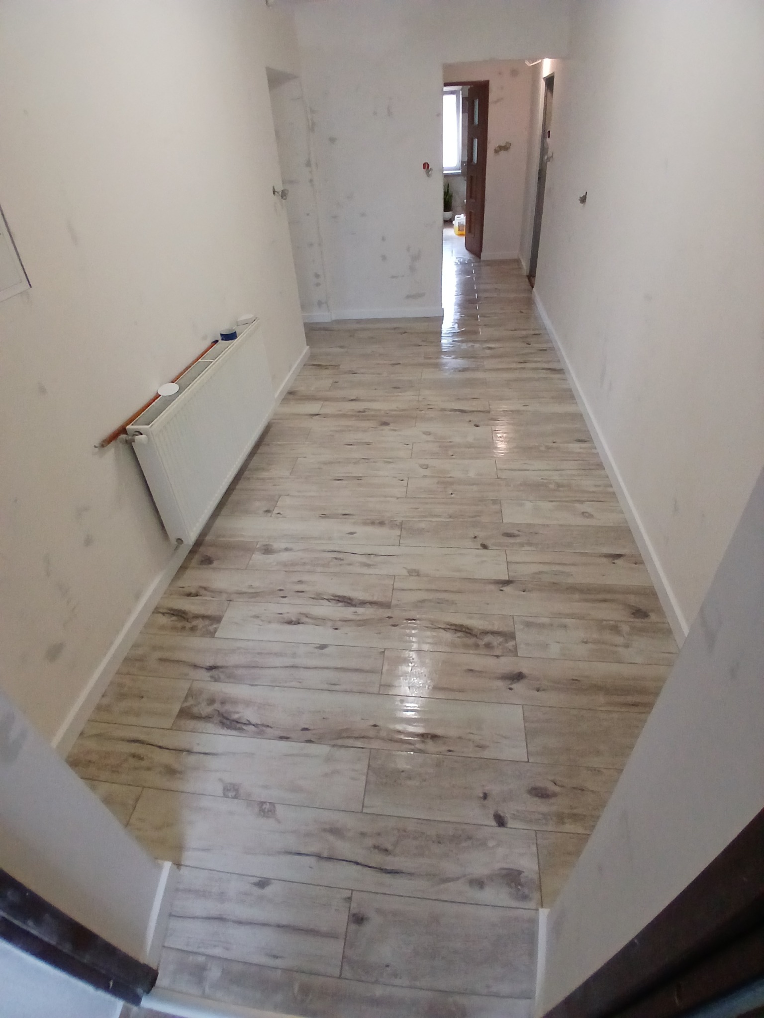 Korytarz po remoncie: świeżo ułożona podłoga z płytek imitujących drewno, gładkie, białe ściany, grzejnik. Widok w głąb mieszkania przez otwarte drzwi.