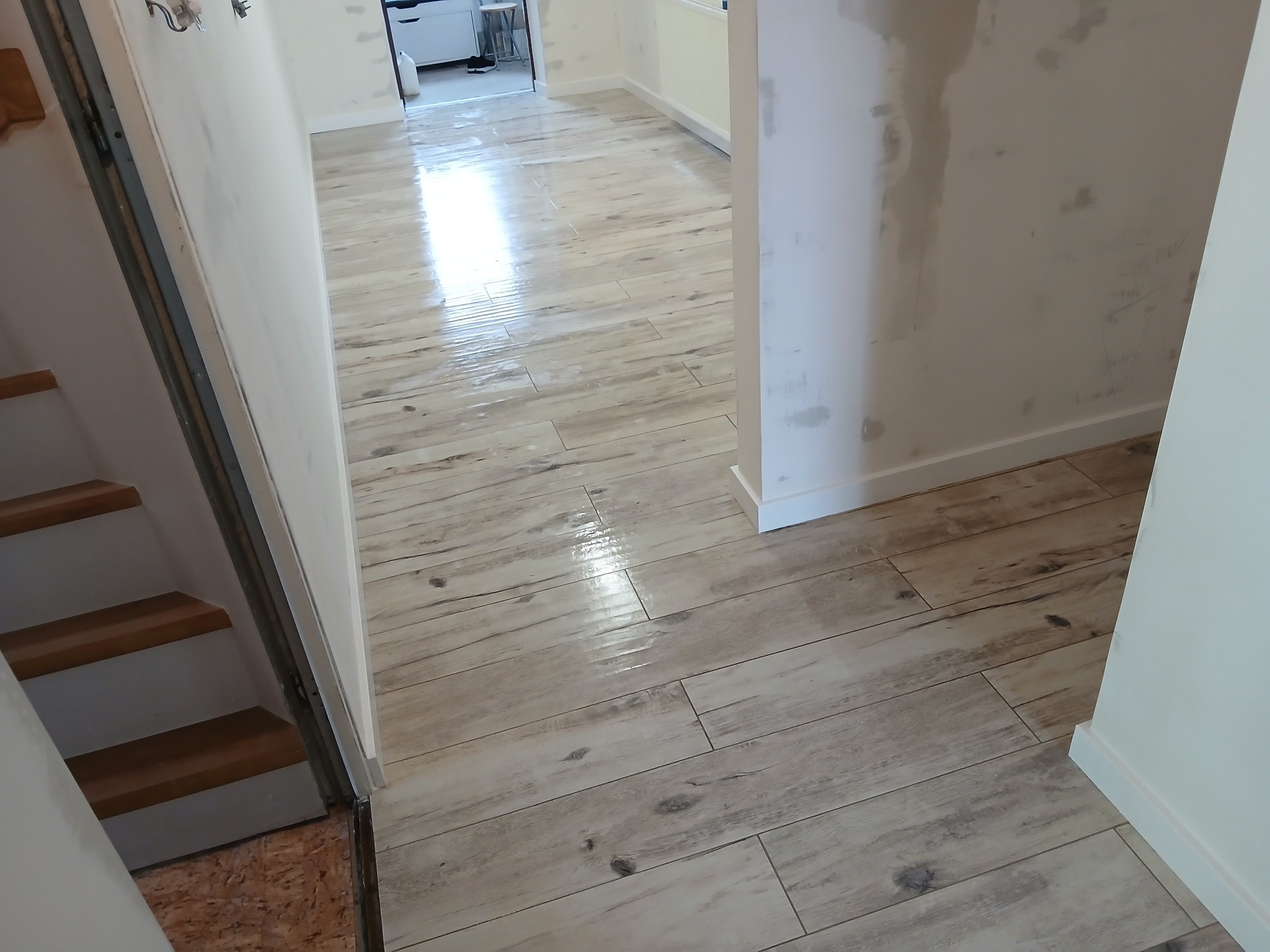Połyskująca, jasna podłoga z płytek imitujących drewno, ułożona w korytarzu. Ściany przygotowane do malowania, widoczne schody. Perspektywa z dołu.