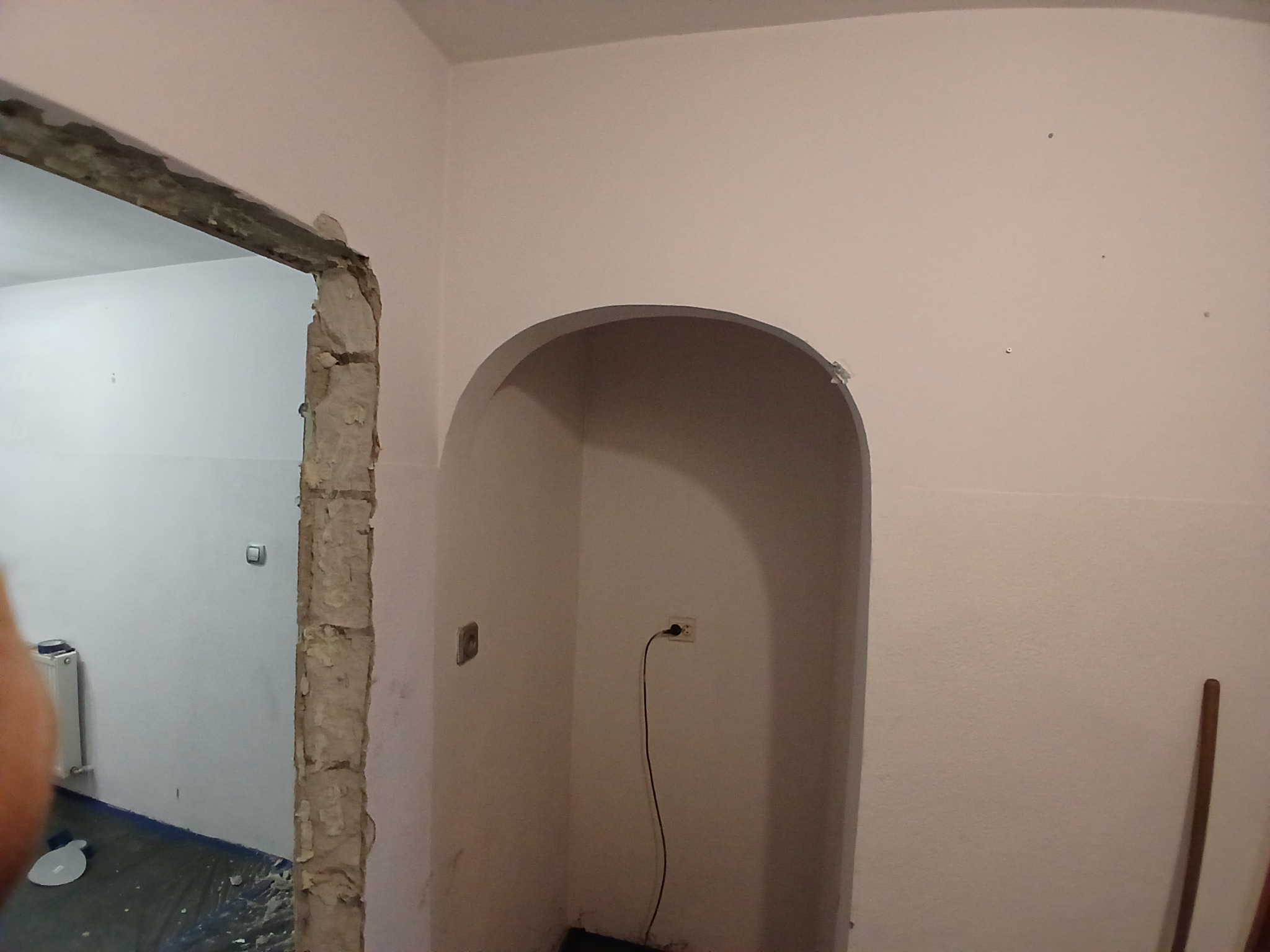 Wnętrze w trakcie prac: surowa ościeżnica, wnęka z łukiem i gniazdkiem. Ściany w jasnym odcieniu, widoczne ślady po remoncie. Podłoga zabezpieczona folią.