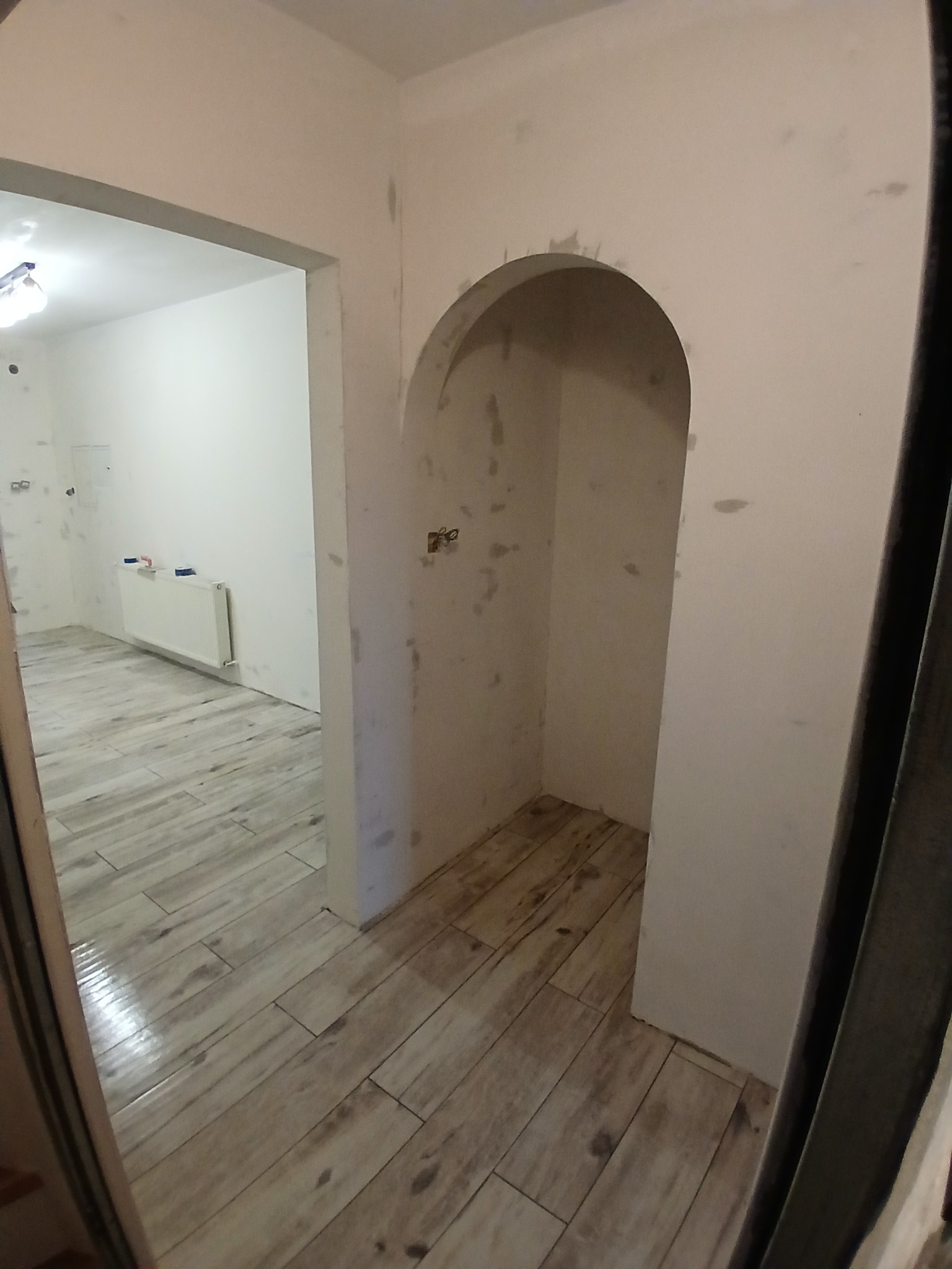 Wnętrze w trakcie prac wykończeniowych: ułożona podłoga z płytek imitujących drewno, ściany przygotowane pod malowanie, wnęka z łukiem. Widok z perspektywy wejścia.