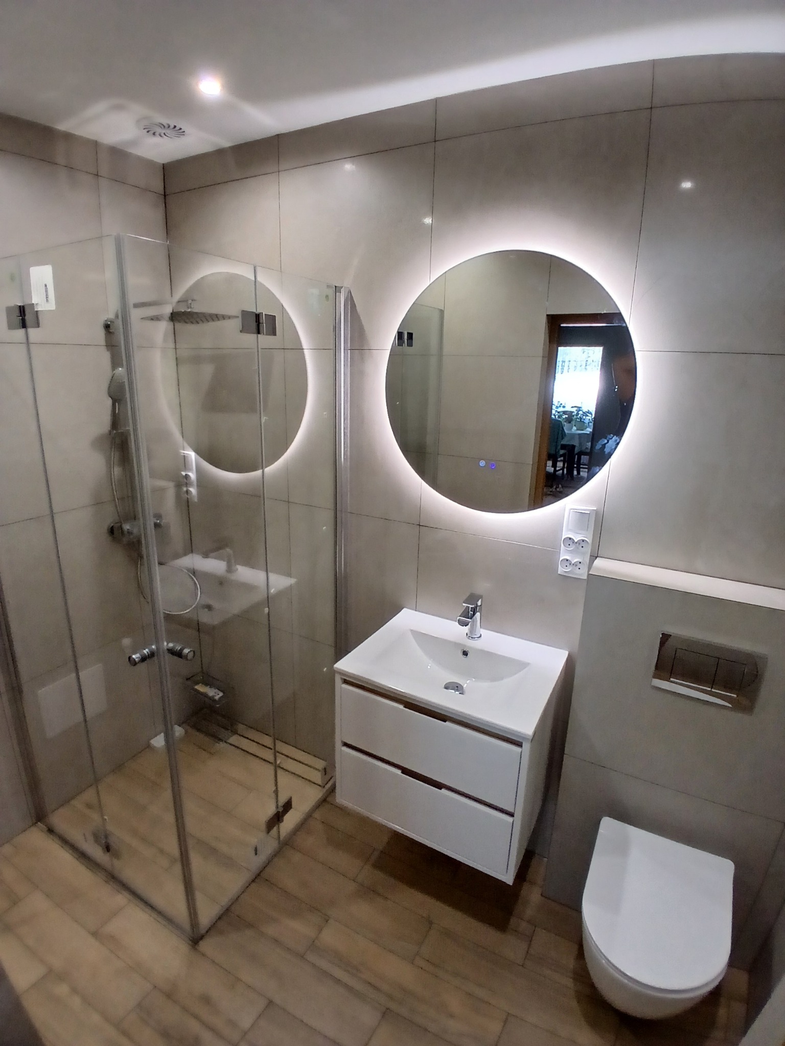 Nowoczesna łazienka z kabiną prysznicową typu walk-in, okrągłym lustrem z podświetleniem LED i podwieszaną szafką z umywalką. Płytki imitujące drewno na podłodze.