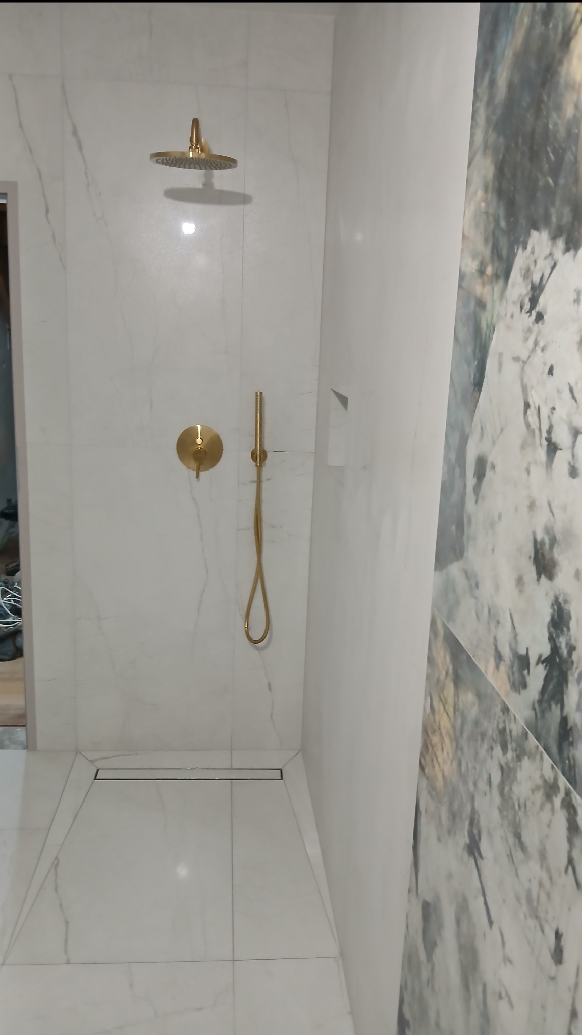 Nowoczesna kabina prysznicowa z odpływem liniowym, wykończona białymi płytkami imitującymi marmur i złotą armaturą. Ściana akcentowa z wzorem przypominającym naturalny kamień.